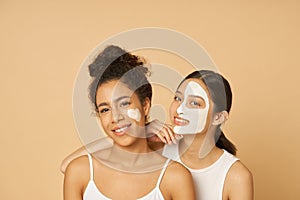 Studio shot of two young women, female friends having fun, smiling at camera while posing with facial masks on isolated