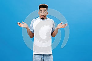 Studio shot of unaware clueless cute african american guy with beard and afro hairstyle shrugging with palms spread