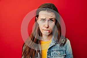 Studio shot of upset young woman purses lower lip, pouting and has displeased look, looks sadly at camera, wears denim jacket,