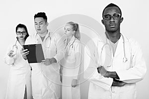 Studio shot of young African man doctor with arms crossed and diverse group of multi ethnic doctors reading on clipboard