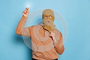 Studio shot of young anonymous man, student wearing balaclava holding bulb isolated on blue background. Concept of