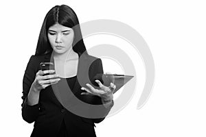 Studio shot of young Asian businesswoman using mobile phone while holding clipboard