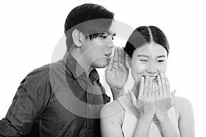 Studio shot of young Asian couple with man whispering to woman laughing