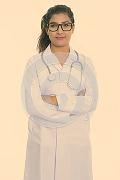 Studio shot of young beautiful Persian woman doctor standing while wearing eyeglasses with arms crossed isolated against