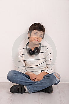 Studio shot of a young boy wearing a white shirt and jeans with headphones siting on the floor by the wall and thinks or sad about
