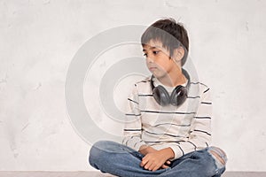 Studio shot of a young boy wearing a white shirt and jeans with headphones siting on the floor by the wall and thinks or sad about