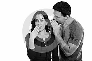 Studio shot of young couple with man whispering to woman looking shocked while covering mouth