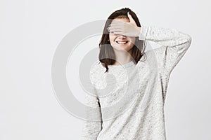 Studio shot of young cute european girl, covering her eyes with hand while smiling cheerfully, being impatient. Mom told