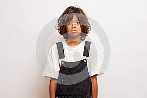 Studio shot of young disappointed teen guy with pouts lips, looks offended, wears denim overall, poses over white wall