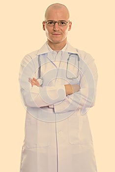 Studio shot of young handsome bald man doctor standing and wearing eyeglasses with arms crossed