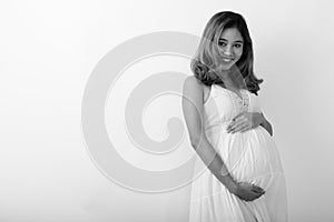 Studio shot of young happy Asian pregnant woman smiling while holding baby against white background