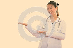 Studio shot of young happy Asian woman doctor smiling while showing something