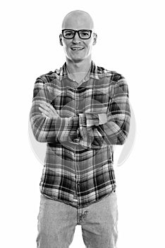 Studio shot of young happy bald man smiling and standing while wearing eyeglasses with arms crossed