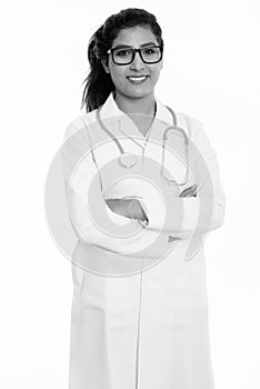 Studio shot of young happy Persian woman doctor smiling and standing while wearing eyeglasses with arms crossed