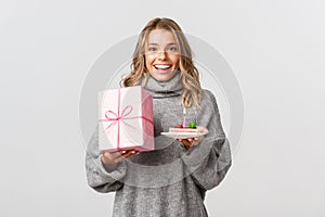 Studio shot of young happy woman holding birthday present and cake, looking cheerful, celebrating her b-day, standing