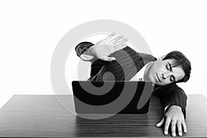 Studio shot of young Persian businessman sleeping while showing stop hand sign with laptop on wooden table