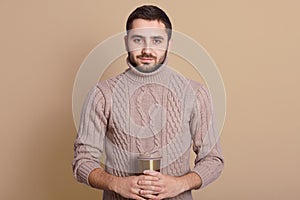 Studio shot of young serious Caucasian man holding coffee cup isolated over beige background, handsome male having thermo mug in