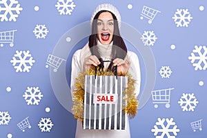 Studio shot of young surprised woman with widely open mouth holding shopping bags in hands with red inscription sale, buys cheep