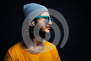 Studio side portrait of young confident man with wireless earphones wearing yellow t-shirt, blue sunglasses and grey hat on black.