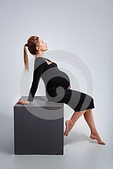 Studio stylish portrait of a pregnant blonde young woman in a black dress posing on a gray background and a cube. The woman is