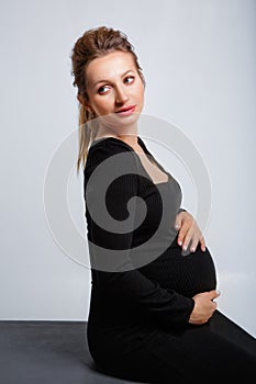 Studio stylish portrait of a pregnant blonde young woman in a black dress posing on a gray background. She sits and gently holds