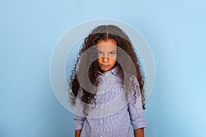 Studio waist up emotional portrait of mulatta frizzy girl wearing blue shirt on a blue background with copy space.. She is