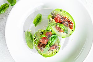 Stuffed avocado halves with sun-dried tomatoes and basil on white plate, top view. Healthy vegan food concept