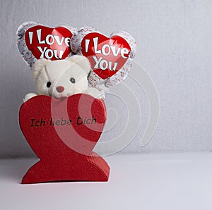 Stuffed bear in white, two balloons with the words `I love you` stuck in a red heart with the words `I love you`