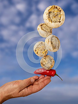 Stuffed mushrooms balancing on a finger and red chilly pepper, blue cloudy sky background. Cooking with top quality fresh products