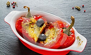 Stuffed red sweet peppers with feta cheese and olives in red and white baking dish on black stone background