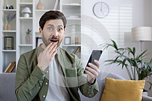 Stunned caucasian guy holding smartphone in hand and cover mouth with hand while looking at camera with amazement