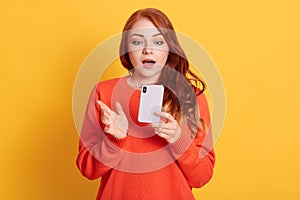 Stunned European woman being in shock, reads terrible news from cellular gadget, has wavy red hair, wears orange sweater, poses