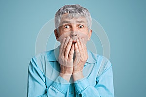 Stunned frightened elderly male model covers mouth with hands, being surprised and scared to hear shocking news, can`t believe in