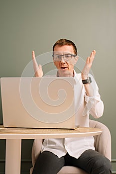 Stunned male furiously raise hands while working on laptop at table. Man in glasses bemused by result, look at monitor.