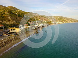 A stunning aerial shot of the blue ocean water along the coastline with homes along the hillside with sandy beaches, blue sky