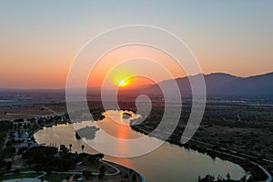 A stunning aerial shot of a sunset over the lake surrounded majestic mountain ranges and lush green trees and grass