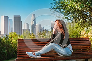 Stunning brunette woman in a denim jacket sits on a park bench with a view of Downtown skyscrapers in Los Angeles