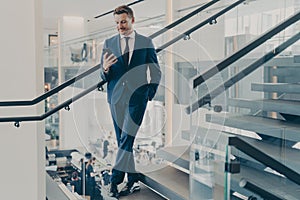 Stunning entrepreneur in formal stylish suit standing on staircase in office lobby with smartphone