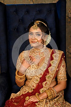 Stunning Indian bride dressed in traditional bridal lehenga with heavy gold jewellery and veil sitting in a chair