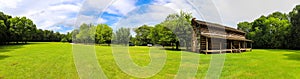 A stunning panoramic shot of a log cabin on a long stretch of lush green grass surrounded by lush green trees with powerful clouds