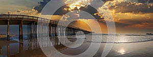This is a stunning panoramic shot of a long winding brown wooden pier at the beach at sunset over the vast blue ocean water