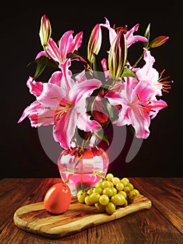 Stunning pink lily bouquet in glass vase on a wooden table and fresh fruit. Still life. Dark background