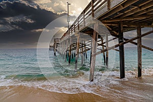 A stunning shot of the Balboa Pier at sunset with powerful clouds, vast blue ocean water and waves crashing into the pier