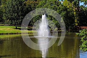 A stunning shot of still silky brown lake with a water fountain in the middle of the lake surrounded by green grass
