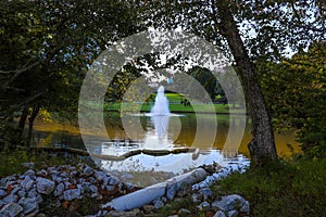 A stunning shot of still silky brown lake with a water fountain in the middle of the lake surrounded by green grass