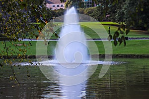 A stunning shot of still silky brown lake with a water fountain in the middle of the lake surrounded by green grass