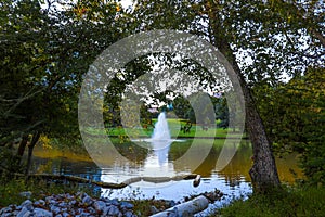 A stunning shot of still silky brown lake with a water fountain in the middle of the lake surrounded by green grass