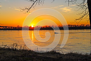 A stunning shot of a white motor boat sailing down the Mississippi river with a gorgeous yellow and red sunset with trees