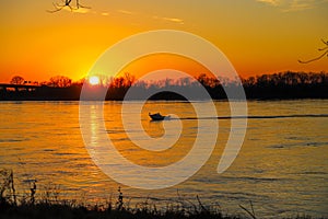 A stunning shot of a white motor boat sailing down the Mississippi river with a gorgeous yellow and red sunset with trees