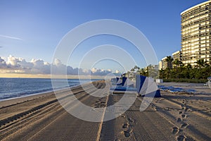 a stunning summer landscape along the beach at sunrise with blue and white lounge chairs, blue ocean water, palm trees, hotels